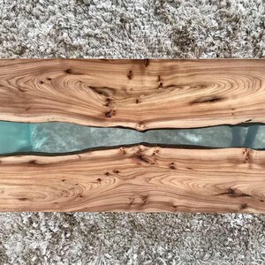 May include: A wooden table top with a natural wood grain finish. The table top has a long, narrow crack filled with clear epoxy resin.