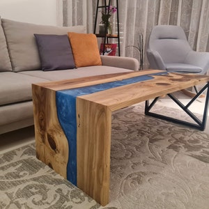 May include: A wooden coffee table with a blue resin river flowing through the center. The table has a black metal frame and is sitting on a patterned rug.