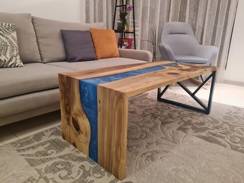 May include: A wooden coffee table with a blue resin river flowing through the center. The table has a black metal frame and is sitting on a patterned rug.