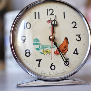 Puede incluir: Un reloj despertador vintage cromado con esfera crema. La esfera del reloj presenta números negros, un segundero rojo y una encantadora ilustración de una gallina y polluelos. Diseño clásico.