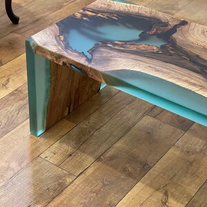 May include: A wooden coffee table with a light blue epoxy resin inlay. The table has a natural wood finish and a unique, organic shape.