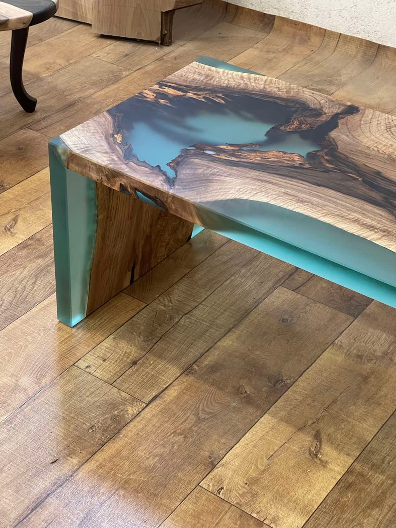 May include: A wooden coffee table with a light blue epoxy resin inlay. The table has a natural wood finish and a unique, organic shape.