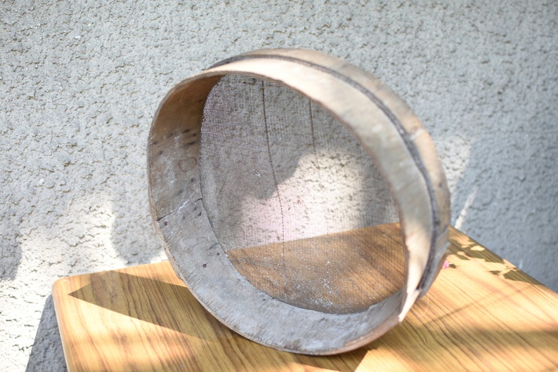 May include: A round wooden sieve with a woven mesh bottom. The sieve is made of light brown wood and has a natural finish. It is sitting on a wooden table.