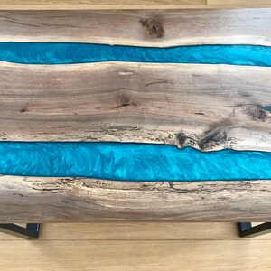 Puede incluir: Una mesa de caf&eacute; de madera con dos incrustaciones de r&iacute;o de resina azul. La mesa tiene un acabado de madera natural y patas de metal negro.