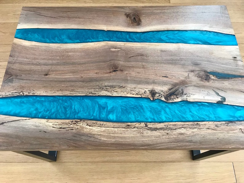 Puede incluir: Una mesa de caf&eacute; de madera con dos incrustaciones de r&iacute;o de resina azul. La mesa tiene un acabado de madera natural y patas de metal negro.