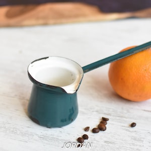 Puede incluir: Una cafetera de esmalte azul verdoso con interior blanco y un mango largo. La cafetera está sobre una superficie de madera blanca con granos de café esparcidos a su alrededor. También hay una naranja en la imagen.