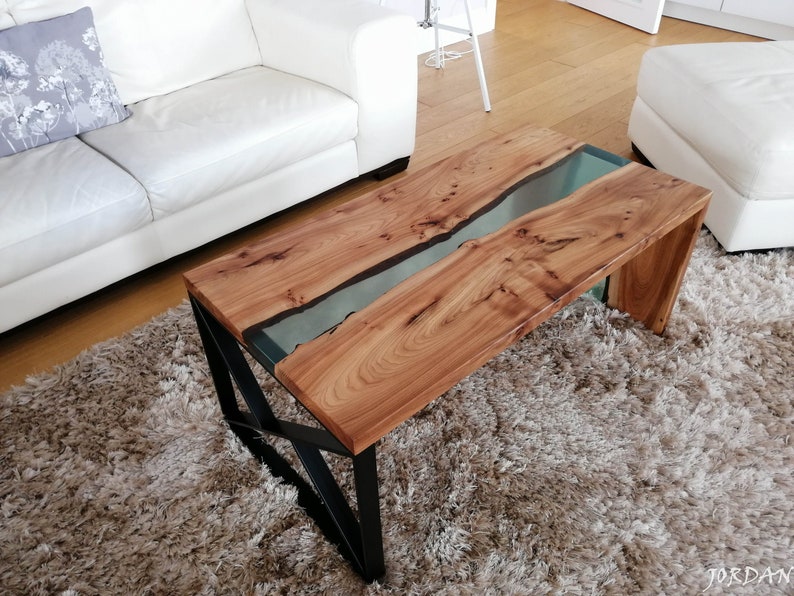 May include: A wooden coffee table with a clear epoxy river running through the center. The table has a black metal frame and is sitting on a fluffy rug.