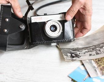 Może przedstawiać: Czarny skórzany futerał na aparat z vintage'owym aparatem w środku. Aparat to czarny i srebrny aparat 35 mm z napisem "OMEHA SUPRA" z przodu. Aparat jest trzymany przez rękę.