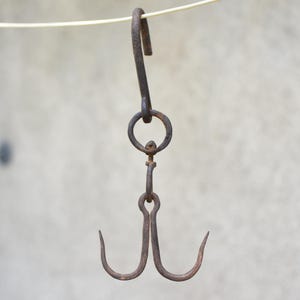 May include: A dark brown, weathered iron meat hook featuring a large top hook connected to a central ring, which attaches to a swivel mechanism and a double-pronged bottom hook. The metal surface shows signs of oxidation and age-related wear.