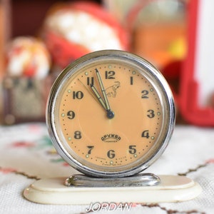 Può includere: Un orologio da tavolo vintage argentato con un quadrante color crema e numeri neri. L'orologio ha una piccola illustrazione di elefante sul quadrante e il testo "Спутник СССР" in scrittura cirillica.
