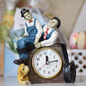 May include: A novelty clock featuring figurines of two men in overalls and bowler hats, with a small dog figurine. The clock face is white with black numbers and gold trim. The time is approximately 12:00.