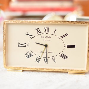 Puede incluir: Un reloj despertador cuadrado de color dorado con marcas de números romanos en la esfera. La esfera del reloj es blanca con manecillas negras y una inscripción negra que dice "Slava 11 Jewels Made in USSR".