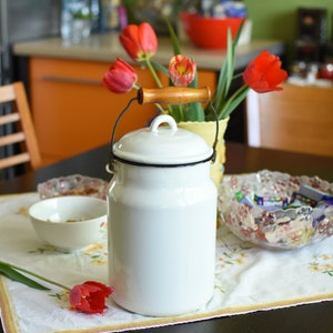 Puede incluir: Cántaro de leche de esmalte blanco con tapa negra y asa de madera. El cántaro está sobre un mantel blanco con estampado floral. Hay un cuenco de nueces y un cuenco de caramelos en la mesa. Hay tulipanes rojos en un jarrón en el fondo.
