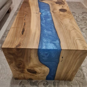 May include: A wooden coffee table with a blue resin river running through the center. The table has a natural wood finish and a square shape.