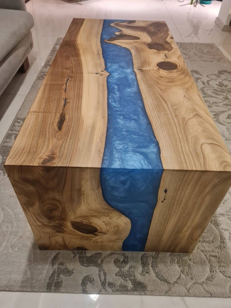 May include: A wooden coffee table with a blue resin river running through the center. The table has a natural wood finish and a square shape.