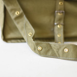 May include: A close-up of a green canvas messenger bag with a strap and brass eyelets.