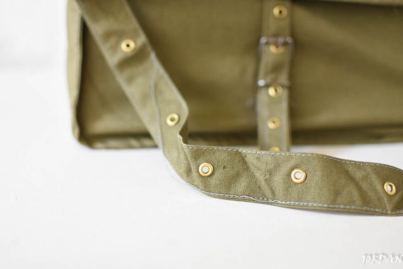 May include: A close-up of a green canvas messenger bag with a strap and brass eyelets.