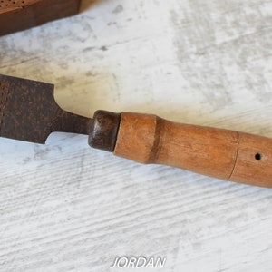 May include: A vintage rasp tool with a wooden handle. The rasp has a dark, textured metal surface and a light brown wooden handle. The tool is designed for shaping wood or other materials. The word "JORDAN" is visible in the image.