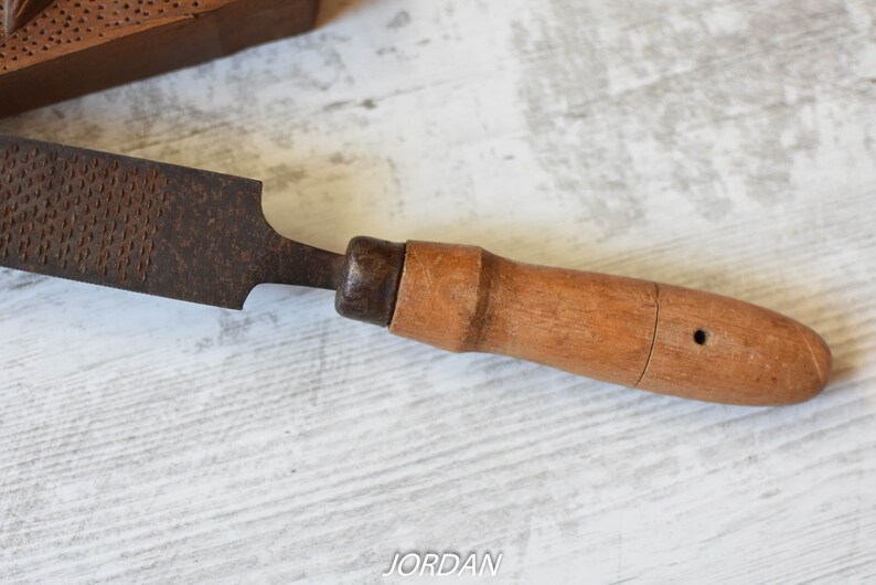 May include: A vintage rasp tool with a wooden handle. The rasp has a dark, textured metal surface and a light brown wooden handle. The tool is designed for shaping wood or other materials. The word "JORDAN" is visible in the image.
