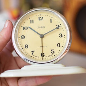 Può includere: Una sveglia Slava vintage color crema con una lunetta color argento. Il quadrante dell'orologio ha numeri e lancette neri, con la scritta "SLAVA" e "11 JEWELS". L'orologio è posto su una base bianca.