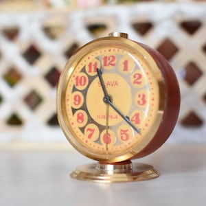 May include: A vintage, round alarm clock with a gold-tone bezel and base. The clock face is cream-colored with red numerals and hands. The clock's body is a dark brown color. The clock reads "SLAVA" on the face.