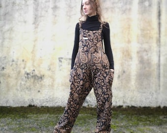 May include: A woman wearing a black turtleneck shirt and black and brown paisley print overalls. The overalls have a wide leg and are cinched at the waist.