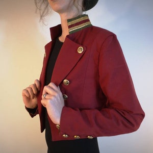 Puede incluir: Una chaqueta roja corta con botones dorados y un ribete verde y dorado alrededor del cuello.