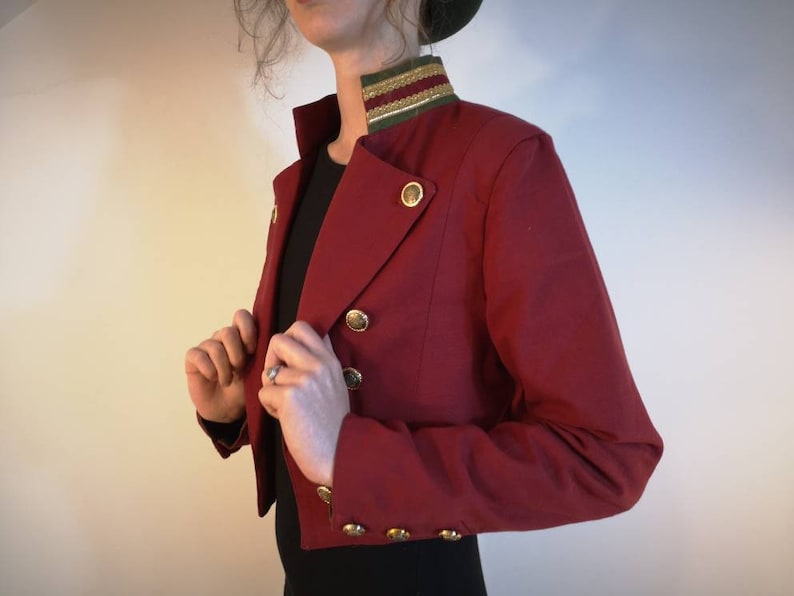Puede incluir: Una chaqueta roja corta con botones dorados y un ribete verde y dorado alrededor del cuello.