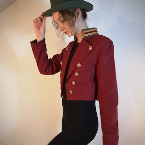 Puede incluir: Una chaqueta roja recortada con botones dorados y ribete verde en el cuello. La chaqueta se lleva sobre una blusa negra y pantalones negros. La persona lleva un sombrero de copa verde.