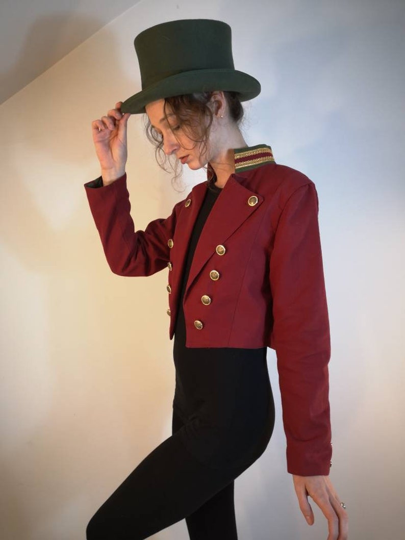 Puede incluir: Una chaqueta roja recortada con botones dorados y ribete verde en el cuello. La chaqueta se lleva sobre una blusa negra y pantalones negros. La persona lleva un sombrero de copa verde.