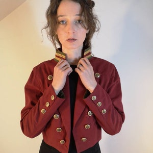 Puede incluir: Una chaqueta roja corta con botones dorados y un ribete verde y dorado alrededor del cuello.