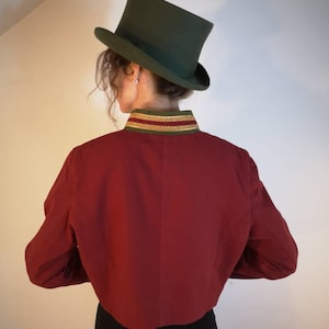 Puede incluir: Una chaqueta roja recortada con ribete dorado y verde alrededor del cuello. La chaqueta tiene mangas largas y la lleva puesta una persona con un sombrero de copa verde.