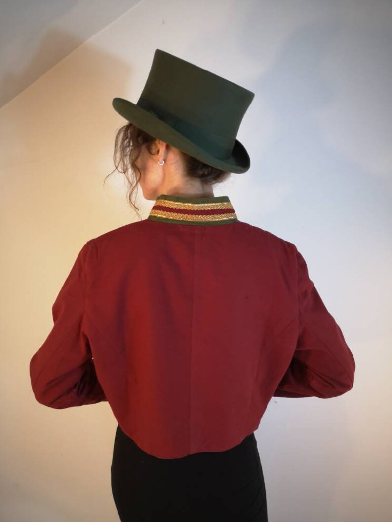 Puede incluir: Una chaqueta roja recortada con ribete dorado y verde alrededor del cuello. La chaqueta tiene mangas largas y la lleva puesta una persona con un sombrero de copa verde.