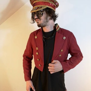 May include: A red cropped jacket with gold buttons and a black shirt underneath. The person is wearing a gold and red sequined hat with a gold band.