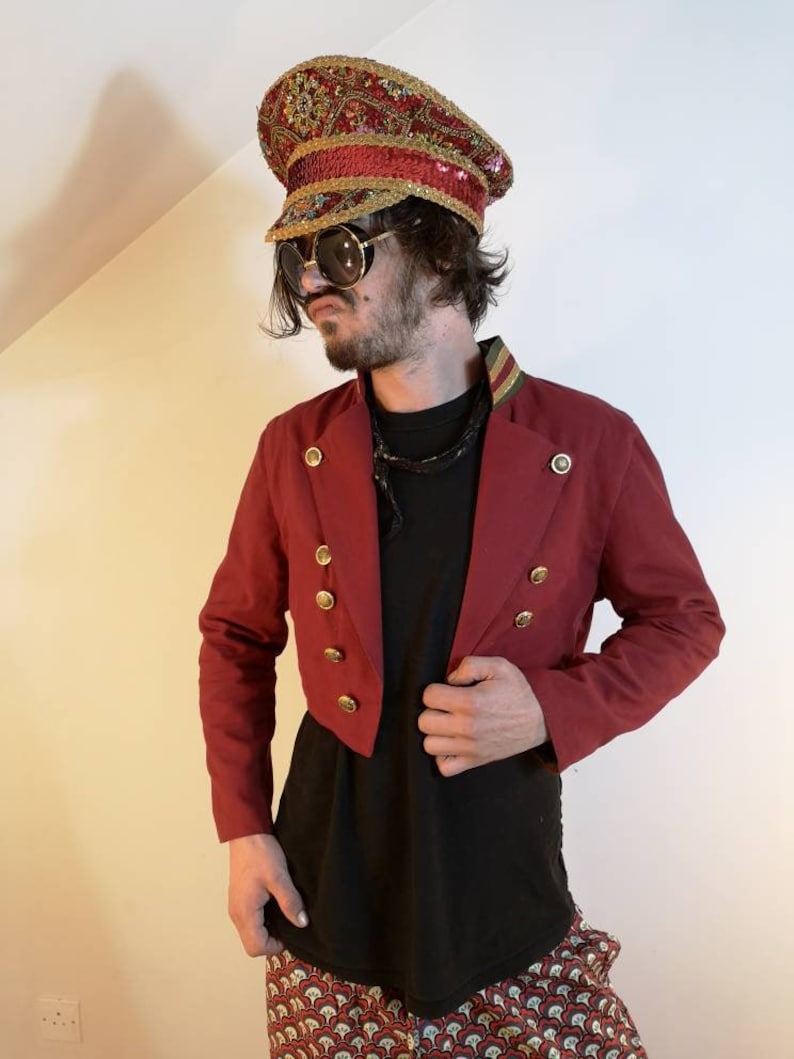 May include: A red cropped jacket with gold buttons and a black shirt underneath. The person is wearing a gold and red sequined hat with a gold band.
