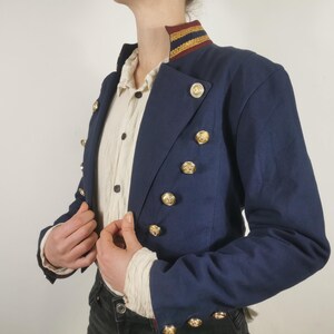 May include: A navy blue cropped jacket with gold buttons and a gold and red trim around the collar.