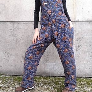 Puede incluir: Pantalón vaquero azul con estampado floral y tirantes marrones. El pantalón tiene un corte holgado y está hecho de una tela suave y cómoda.