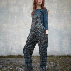 May include: A woman wearing a blue and brown patterned jumpsuit with a teal long-sleeved top. She is standing in front of a grey concrete wall.
