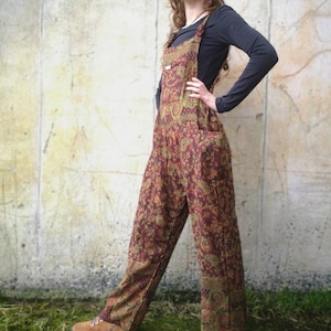 May include: A woman wearing a pair of brown and red paisley print dungarees with a black long-sleeved top. She is standing in front of a concrete wall.