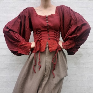 Op de afbeelding: Een bordeauxrode blouse met een vierkante halslijn en lange, volumineuze mouwen. Het lijfje heeft een gesnoerd korsetontwerp en knopen. De mouwen hebben kanten afwerking aan de manchetten. De blouse wordt gecombineerd met een beige broek.