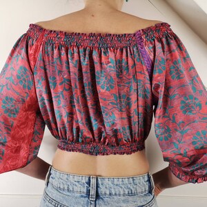 May include: A red and teal floral print off-the-shoulder crop top with a gathered elastic neckline and long sleeves. The top is made of a soft, silky fabric.