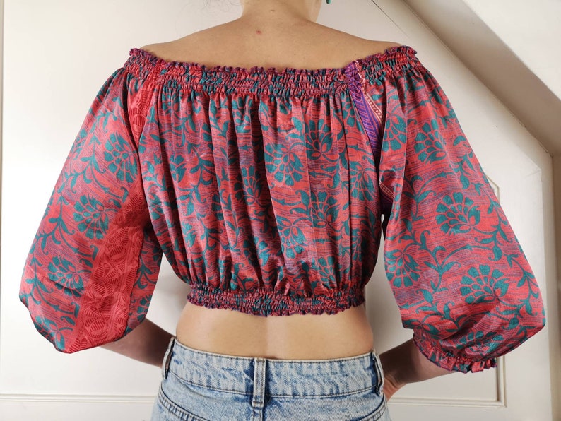 May include: A red and teal floral print off-the-shoulder crop top with a gathered elastic neckline and long sleeves. The top is made of a soft, silky fabric.