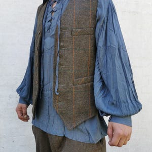 Könnte beinhalten: Ein Outfit mit mehreren Schichten, bestehend aus einem blauen Hemd mit Schnürhalsausschnitt und einer braunen Tweedweste mit orangefarbenen Streifen. Abgerundet wird das Outfit durch eine braune karierte Hose, die einen rustikalen, Vintage-Look erzeugt.