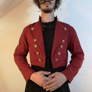 May include: A red cropped jacket with gold buttons and a black collar. The jacket has a fitted silhouette and a short length.