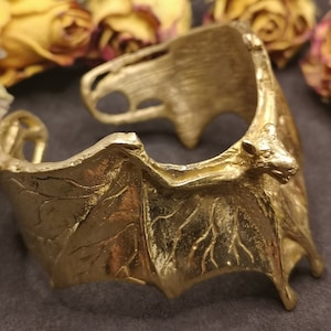 Op de afbeelding: Een goudkleurige manchetarmband met een vleermuisontwerp. De armband wikkelt zich om de pols en heeft gedetailleerde vleugels en een vleermuiskop. Het metaal heeft een getextureerd uiterlijk en het algehele ontwerp is gotisch en uniek.