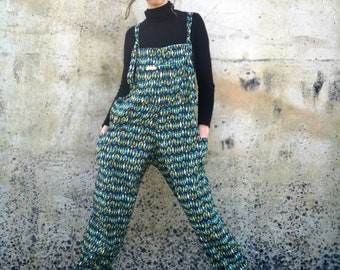 May include: A person wearing a black turtleneck shirt and blue and green patterned overalls. The overalls have large pockets on the sides. The person is standing in front of a white concrete wall.