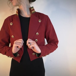 Puede incluir: Una chaqueta roja corta con botones dorados y un cuello a rayas verde y dorado.