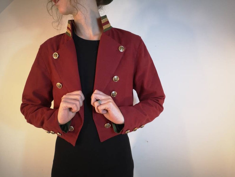 Puede incluir: Una chaqueta roja corta con botones dorados y un cuello a rayas verde y dorado.