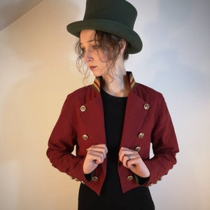 Puede incluir: Una chaqueta roja recortada con botones dorados y un ribete verde y dorado alrededor del cuello. La chaqueta se lleva sobre una camisa negra y la persona lleva un sombrero de copa verde.