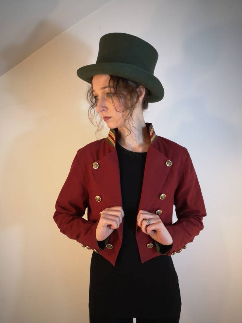 Puede incluir: Una chaqueta roja recortada con botones dorados y un ribete verde y dorado alrededor del cuello. La chaqueta se lleva sobre una camisa negra y la persona lleva un sombrero de copa verde.
